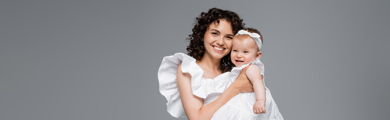 Happy mother in white dress hugging baby daughter isolated on grey, banner.