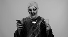 Sinister Man With Horrible Scary Halloween Zombie Make-up Using Credit Bank Cards And Mobile Phone, Transferring Money Purchases Online Shopping. Dead Guy With Wounded Bloody Scars Face, Gray Room