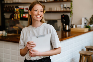 Wall Mural - Blonde white woman smiling and holding clipboard while working in cafe