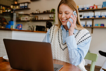 Wall Mural - Blonde woman talking on cellphone while working with laptop at cafe