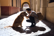 Young Hispanic Man With Beard And Sunglasses Crouching With His Dog While Hugging Him On A Sunny Street At Sunset Casting His Shadow On The Ground. Concept Animals, Dogs, Love, Pets, Golden.