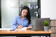 © wichayada - Charming asian businesswoman working on laptop and taking note while sitting at the table in office.