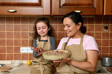 Wall Mural - Beautiful young dark-haired multi-ethnic woman, a loving mother smiling cheerfully, standing near her cute little girl holding her first baked little cherry pie, sitting on the kitchen countertop