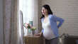 © PRPicturesProduction - asian woman approaching to the window is massaging herself while feeling sudden lower back pain during pregnancy in the morning at home.