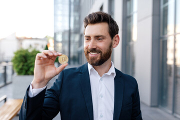 Wall Mural - Glad confident attractive young caucasian guy with beard in suit shows bitcoin coin outdoor