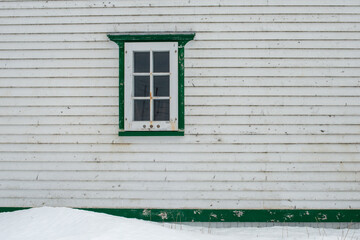 The exterior outside wall of a stark white heritage wooden building with a single window. The country style vintage house has narrow white clapboard siding and vibrant green trim with a white snowbank