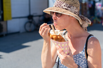 Attractive woman wearing sunglasses and straw hat, in a light summer dress, eating ice cream off a bubble waffle on the street. Summer holiday, vacation, leisure.