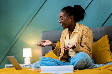 Wall Mural - Young African American woman sitting in bed in the bedroom studying using laptop.