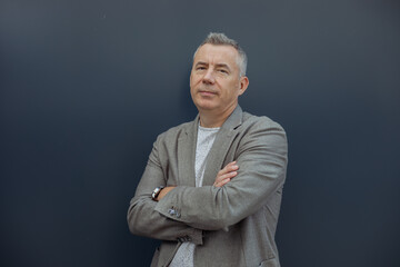 Portrait of elegant aged gray haired businessman enjoying and standing with crossed arms in stylish suit on gray background. Urban lifestyle, entrepreneur, well dressed man, positive emotion