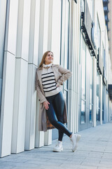 Wall Mural - A cute young woman in a beige raincoat and autumn leather boots walks down the street. Autumn warm image. Women's fashion