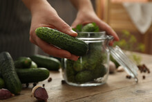 Woman Putting Cucumber Into Glass Jar At Wooden Kitchen Table, Closeup. Pickling Vegetables