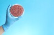Scientist holding Petri dish with minced lab grown meat on light blue background, closeup. Space for text