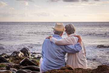 Sticker - Rear view of relaxed caucasian senior couple sitting on the pebble beach at sunset light admiring horizon over water. Two gray haired elderly people hugging with love