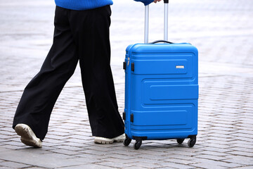 Wall Mural - Slim girl in black pants walking with blue suitcase on wheels. Female legs and luggage on the street, travel concept