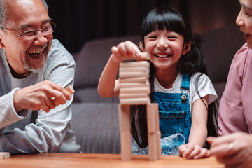 Asian happy family of little daughter play toy wooden block in home. Grandparents and young woman loving couple parent having fun play stack tower wood block game at night in living room.