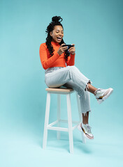 Portrait of an excited young woman sitting on a stool and playing games on mobile phone isolated over blue background.