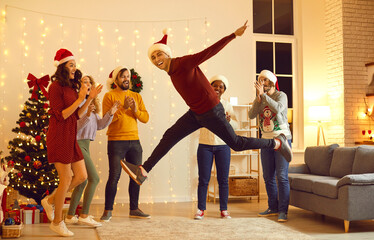 Overjoyed multiethnic friends in Santa hats have fun celebrate New Year in decorated home near fir-tree. Happy diverse young people enjoy Christmas celebration together. Winter holidays concept.