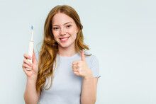 Young Caucasian Woman Holding Electric Toothbrush Isolated On Blue Background Smiling And Raising Thumb Up