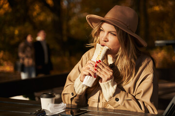 a beautiful young woman sits in an outdoor cafe at a wooden table in an autumn park, and eats kebab or shawarma