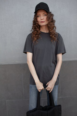 Wall Mural - A beautiful girl is wearing a grey t-shirt,black bag,  cloth cap and jeans posing against the background of a wall. fashion outfit for the city, minimalism urban style clothing