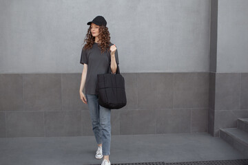 Wall Mural - A beautiful girl is wearing a grey t-shirt,black bag,  cloth cap and jeans posing against the background of a wall. fashion outfit for the city, minimalism urban style clothing