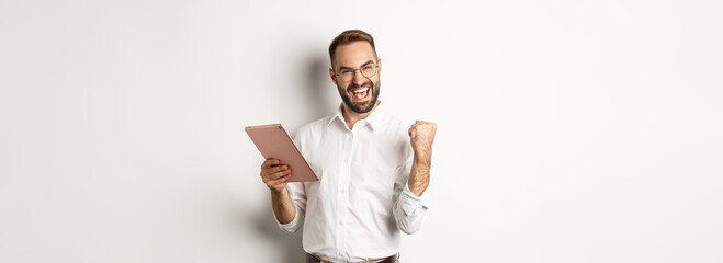 Wall Mural - Successful businessman rejoicing on winning online, reading on digital tablet and making fist pump, triumphing, standing over white background