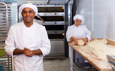 Wall Mural - Portrait of confident hispanic bakery owner in white uniform posing against busy workers background