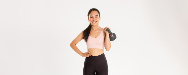 Wall Mural - Sport, wellbeing and active lifestyle concept. Smiling strong and slim asian fitness girl, female athlete holding kettlebell and looking carefree, gaining muscles at gym, standing white background