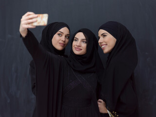 Group of young muslim women in fashionable dress with hijab using smartphone while taking selfie picture in front of black background 