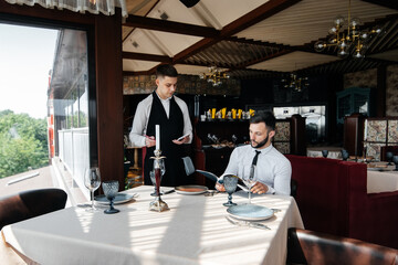 Wall Mural - A young businessman in a fine restaurant examines the menu and makes an order to a young waiter in a stylish apron. Customer service. Table service in the restaurant.