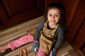 Wall Mural - View from above to a pretty child girl, little baker confectioner in a beige chef apron and kitchen mittens, sitting on the floor and leaning against wooden kitchen cupboard, sweetly smiling to camera