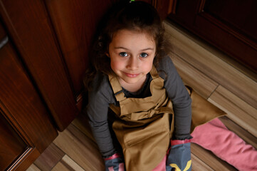 Wall Mural - Overhead view of a Caucasian child, baby girl, cute little baker confectioner, chef pastry in a beige apron and mittens, sitting barefoot on the kitchen floor and cutely smiling looking at camera