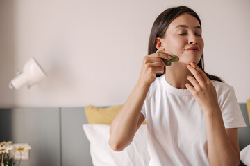 Beautiful young caucasian woman enjoys morning routine with face massager at home. Brunette hair girl sitting on bed in morning. Concept scraper, massage.