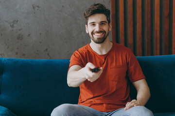 Wall Mural - Young happy man wears red t-shirt hold TV remote controller watch film switch channels sit on blue sofa couch stay at home hotel flat rest relax spend free spare time in living room indoors grey wall.