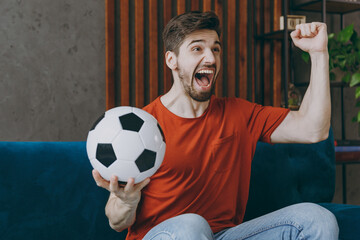 Wall Mural - Young excited fan man in red t-shirt cheer up support football team sport team hold soccer ball watch tv live stream do winner gesture sit on sofa stay home rest spend free spare time in room indoor.