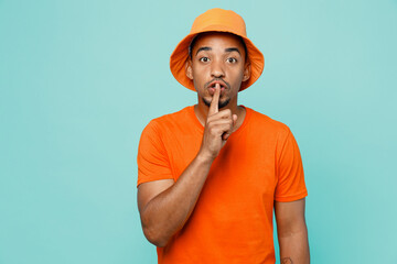 Wall Mural - Young secret man of African American ethnicity wear orange t-shirt hat say hush be quiet with finger on lips shhh gesture isolated on plain pastel light blue cyan background. People lifestyle concept.