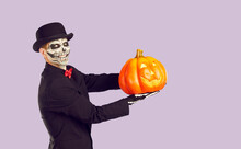 Portrait Of Cheerful Man With Traditional Halloween Skeleton Make-up Holding Glowing Pumpkin In His Palm. Man In Black Suit And Hat Is Holding Pumpkin And Smiling At Camera On Light Lilac Background.