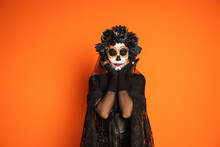 Woman With Spooky Halloween Makeup Wearing Black Costume And Wreath Standing With Closed Eyes Isolated On Orange.