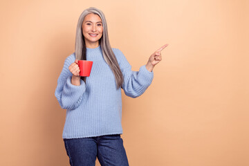 Wall Mural - Photo of funny pretty retired woman wear blue sweater drinking coffee pointing empty space isolated beige color background