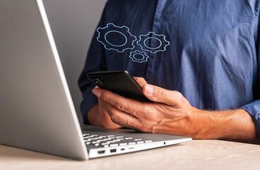 Poster - Man holding phone with virtual gears while working on laptop. Mobile settings or strategy, process organization, business idea concept.