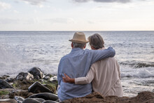 Rear View Of Relaxed Caucasian Senior Couple Sitting On The Pebble Beach At Sunset Light Admiring Horizon Over Water. Two Gray Haired Elderly People Hugging With Love