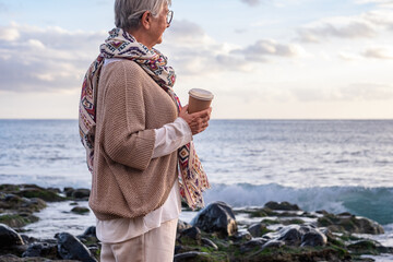 Smiling caucasian senior woman standing at the beach holding coffee cup looking at the horizon over sea. Elderly lady at sunset enjoying view and nature