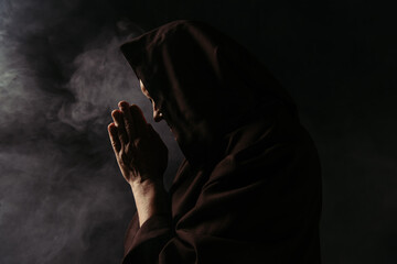 Canvas Print - side view of priest with face obscured by hood praying on black with smoke.