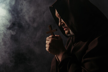 Poster - side view of monk in robe holding holy cross while praying on black with smoke.