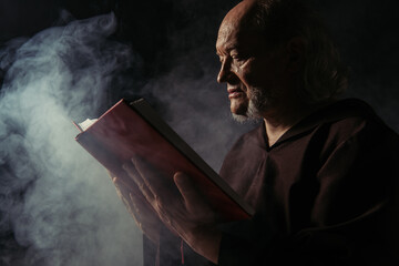 Wall Mural - side view of priest in black robe reading holy bible on dark background with smoke.