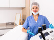 Portrait of positive female doctor cosmetologist wearing gloves and blue overall showing equipment of clinic before procedure