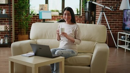 Poster - Joyful asian woman sitting in living room working from home while smiling at camera. Happy young adult student inside modern apartment having laptop on table while waiting for online class to start.