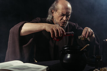 Wall Mural - serious alchemist pouring liquid into pot near magic cookbook isolated on black.