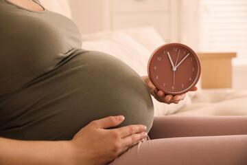 Wall Mural - Young pregnant woman holding clock near her belly at home, closeup. Time to give birth