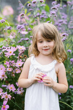 Little Toddler Girl In White Dress Playing In The Grass By Flowers